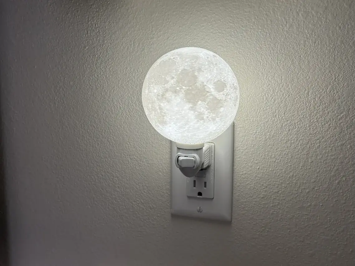 The small moon night light lit in a wall outlet at night, a complete glowing sphere above its white swivelling fixture