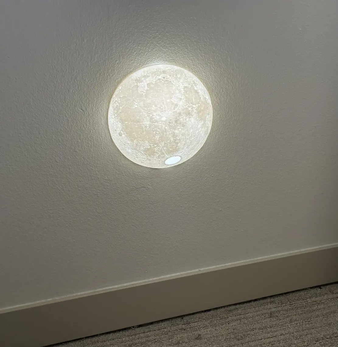 The lit moon night light on a hallway wall above the baseboard, the round switch cutout showing as a small pale oval at the bottom of its face